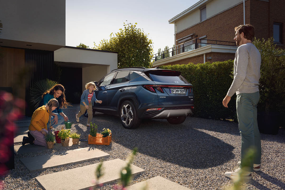 zu sehen ist ein blauer Hyundai TUCSON vor der Garage, nebenan pflanzt eine Familie Blumen